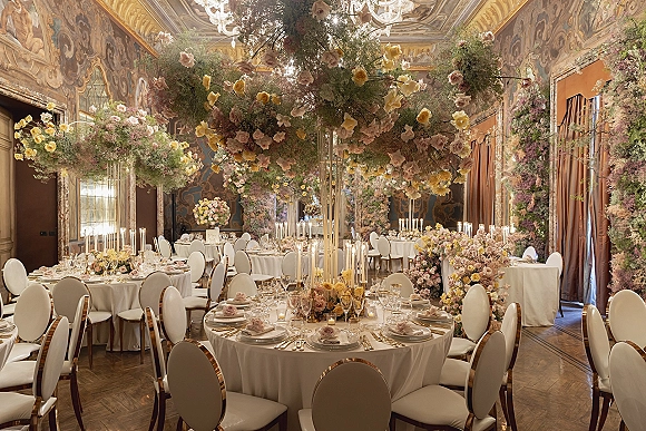 Reception tablescape with hanging floral installation and tall taper candles on round white linen tables beneath chandeliers and ceiling murals