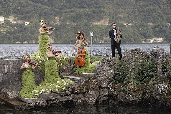 Wedding musicians perform lakeside wedding string quartet music in green ruffled dresses with floral hairpieces, violins and cello on stone steps