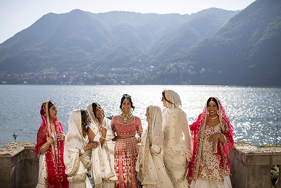Bridesmaid group portrait of South Asian bridesmaids in embroidered lehengas with gold jewelry, laughing on a lakefront terrace with mountains behind