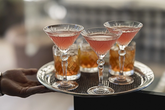 Wedding cocktails on a serving tray with coupe glasses and rocks glasses over ice on a bar counter in an indoor reception space