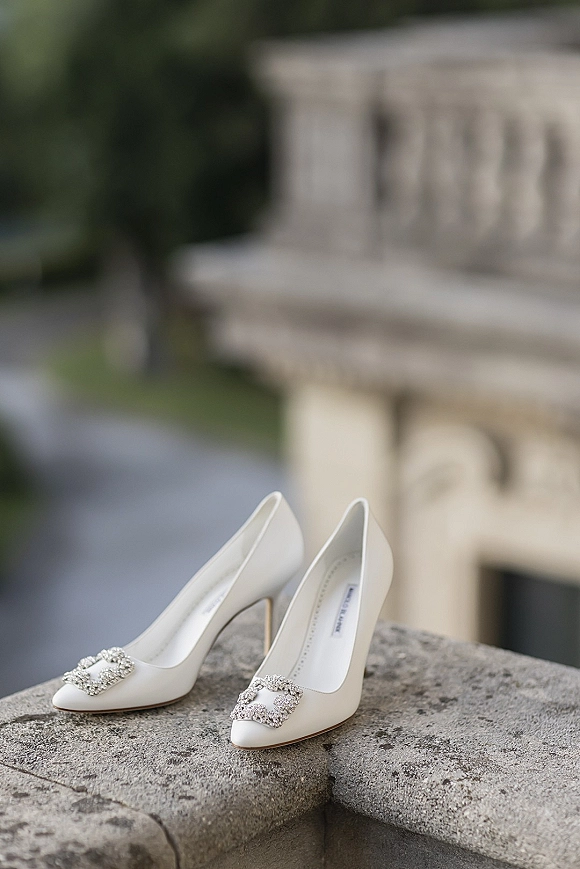 Bridal shoes in ivory wedding heels with jeweled buckles resting on a stone ledge beside greenery and a stone balustrade