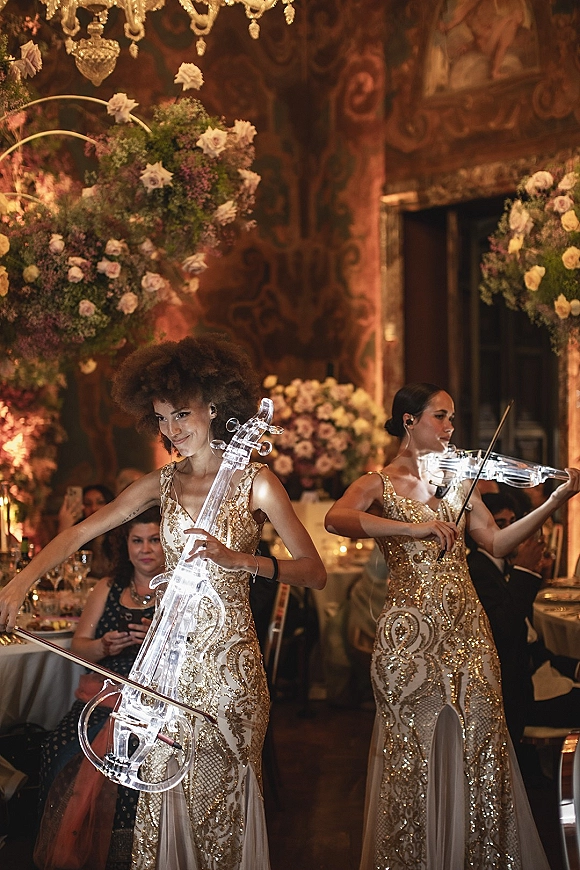 Wedding reception entertainment with wedding string performers playing electric cello and violin in gold sequin gowns beneath chandelier in ornate ballroom