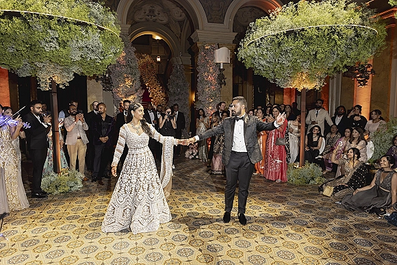 Wedding first dance as bride in a lehenga and groom in tuxedo twirl under a floral ceiling in an arched hall with guests watching