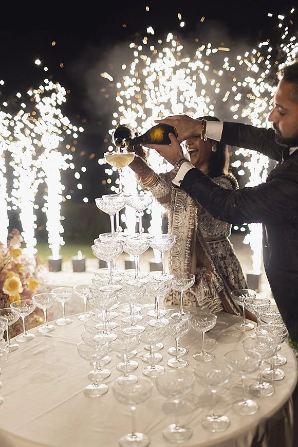 Champagne tower moment as newlyweds pour from a bottle into coupe glasses, sparklers flaring under the night sky on an outdoor lawn