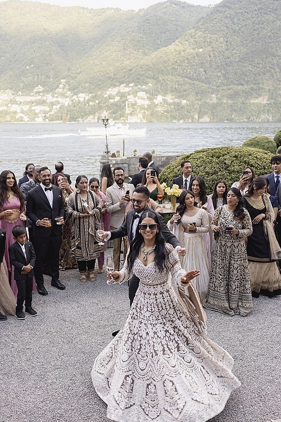 Wedding reception dancing as bride in lehenga and groom in black tuxedo celebrate on a lakeside stone terrace with mountain backdrop