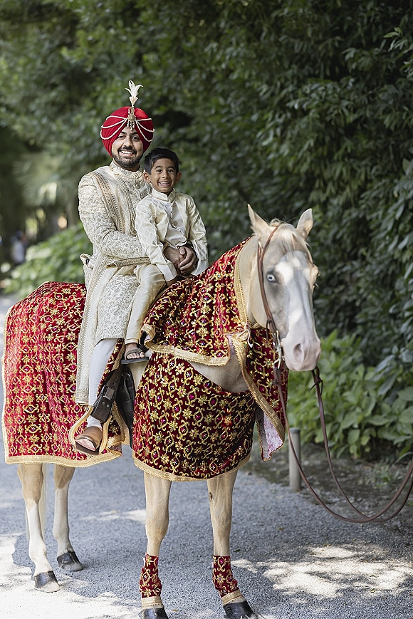 Groom baraat portrait of the groom on horse, wearing an embroidered sherwani and red turban with feather brooch on a tree-lined path