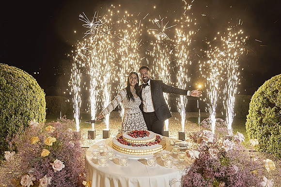 Wedding cake cutting with sparkler cake cutting fountains as bride in long-sleeve gown and groom in tux slice berry cake at night garden table