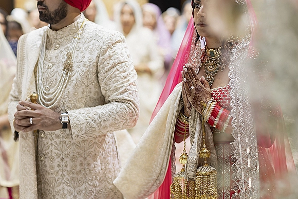 Ceremony moment at a Sikh wedding ceremony with bride’s mehndi hands in prayer beside groom in embroidered sherwani, guests in soft bokeh lights