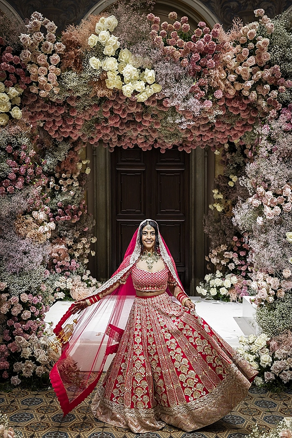 Bridal portrait of an Indian bride in a red lehenga with dupatta veil and gold jewelry, seated by an ornate indoor doorway and floral arch