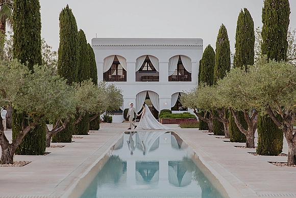 Couple portrait of bride and groom walking hand in hand, her long veil trailing by a reflecting pool at a white villa garden walkway