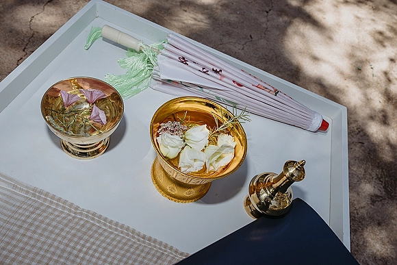 Ceremony water bowl with floating flower petals and herbs, set in gold pedestal bowls on a white tray in dappled sunlight