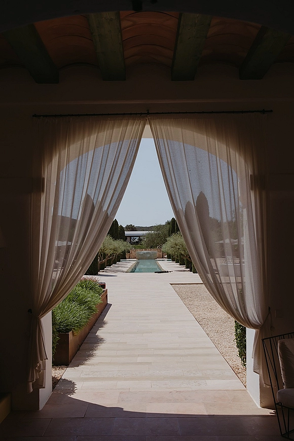 Wedding venue entrance with draped wedding entrance sheer curtains on pergola beams, stone walkway by a pool in a courtyard of trees