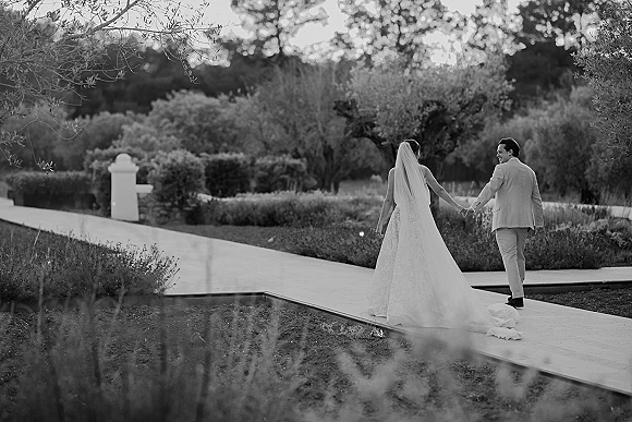 Couple portrait of bride and groom holding hands, walking away on a garden path, her long veil and dress train flowing behind