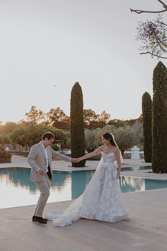 Couple portrait of bride and groom holding hands, bride in veil, strolling poolside on a stone patio in warm sunset light