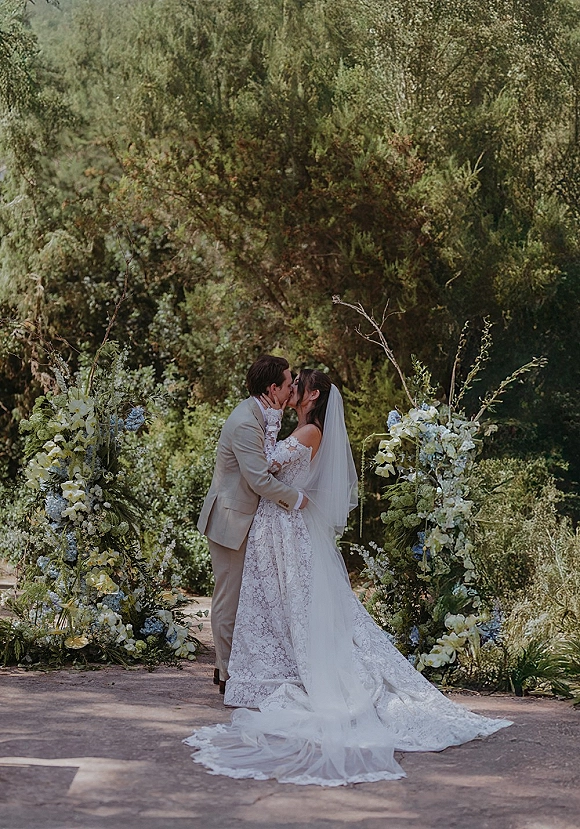 Wedding kiss portrait of bride and groom kissing beneath a hydrangea-filled garden arch, her veil and lace train flowing on a stone patio