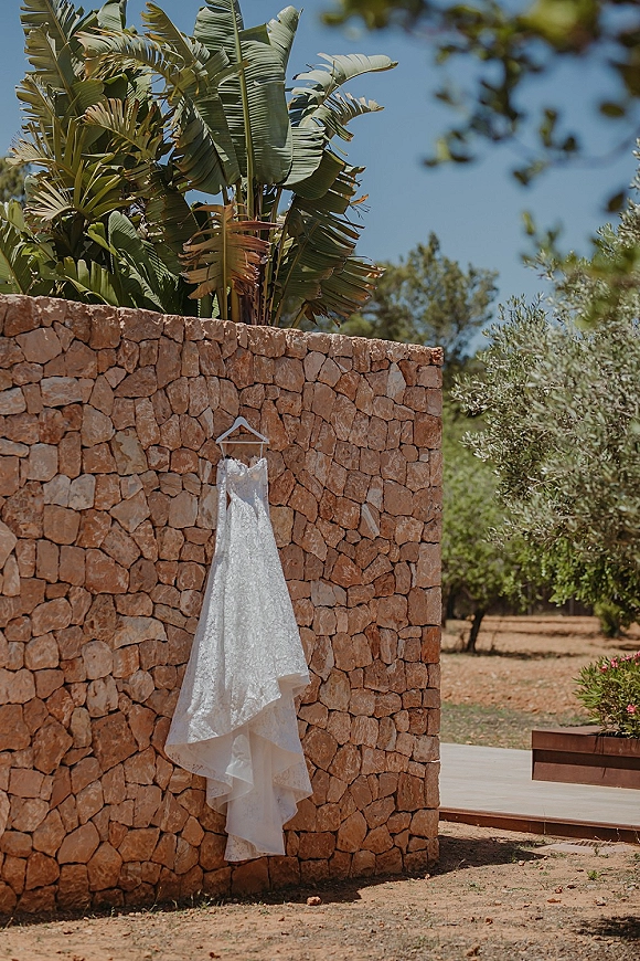 Wedding dress on a hanger, lace wedding dress details catching natural light against a stone wall with palm leaves and blue sky