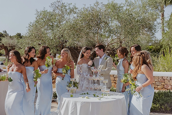 Wedding party toast around a champagne tower, bride in lace veil and groom in beige suit cheering in an olive grove patio setting