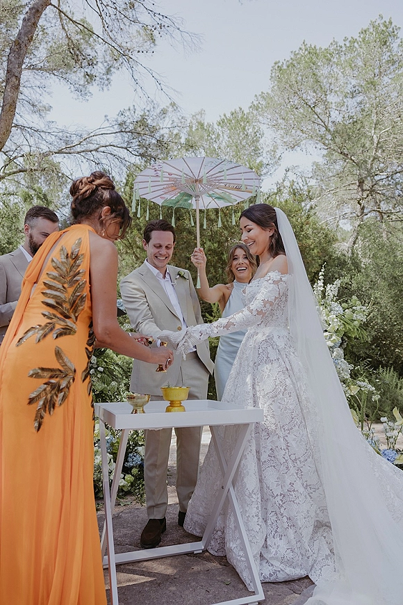 Ceremony moment at an outdoor wedding ceremony as the bride and groom stand by a white table while guests pop confetti in a garden path
