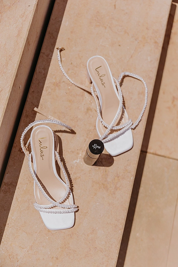 Bridal shoes with pearl wedding heels straps and square toes beside a small perfume bottle on sunlit stone steps with shadows
