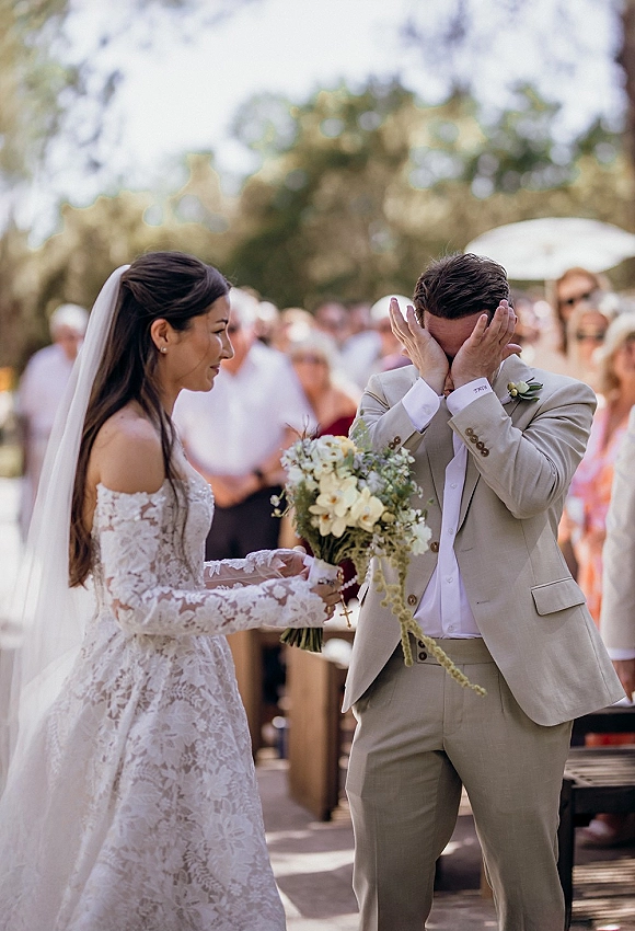 First look moment as groom covers his face, moved by bride in lace wedding dress holding bouquet on a sunlit outdoor aisle