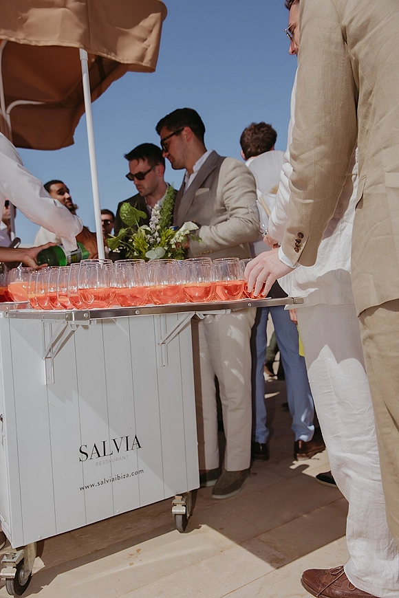 Wedding cocktail hour with champagne coupe tower on a cocktail cart as guests in linen suits mingle under a patio umbrella on a sunny deck
