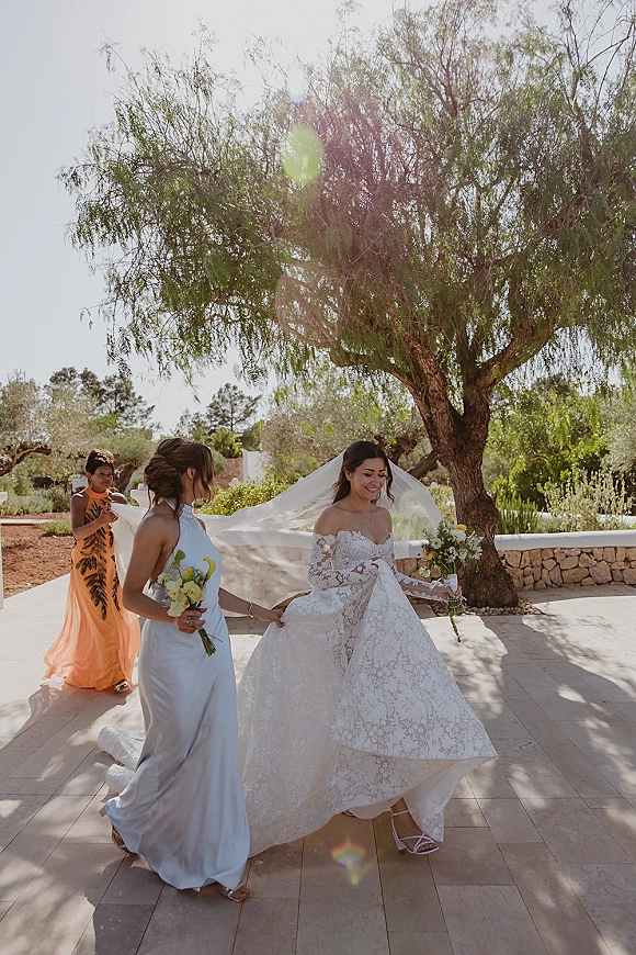 Bride portrait of a smiling bride holding bouquet in an off-the-shoulder lace wedding dress with veil, on a sunlit garden patio by stone wall