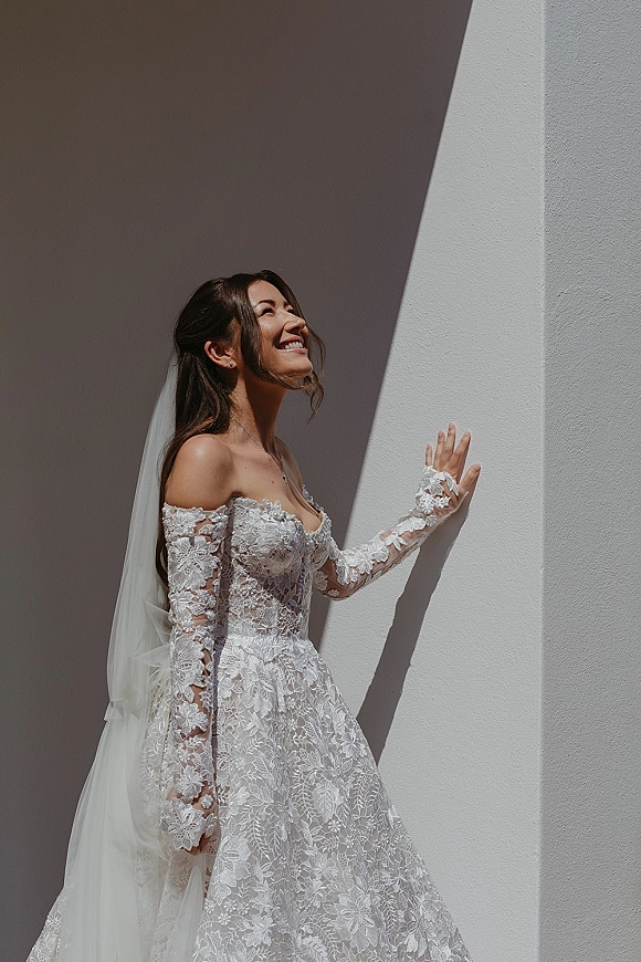 Bridal portrait of a smiling bride in an off the shoulder lace wedding dress with cathedral veil, posing by a white wall in hard sunlight shadows