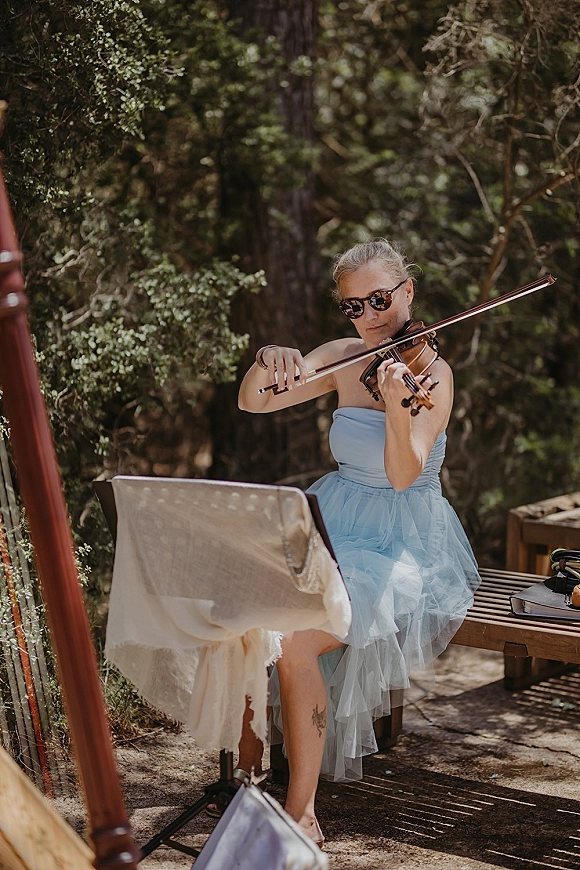Wedding musician wedding violinist in sunglasses plays violin beside a music stand with sheet music on a wooded deck outdoors