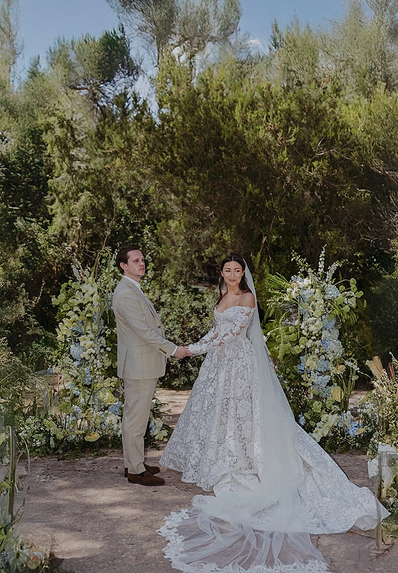 Ceremony moment as bride in lace dress and veil holds hands with groom in beige suit on a garden aisle with florals and greenery