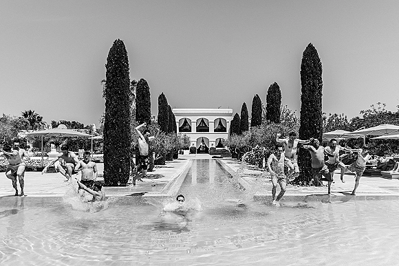 Groomsmen pool photo of friends jumping into outdoor pool in swim trunks, splashing by a villa pool deck with umbrellas and tall trees