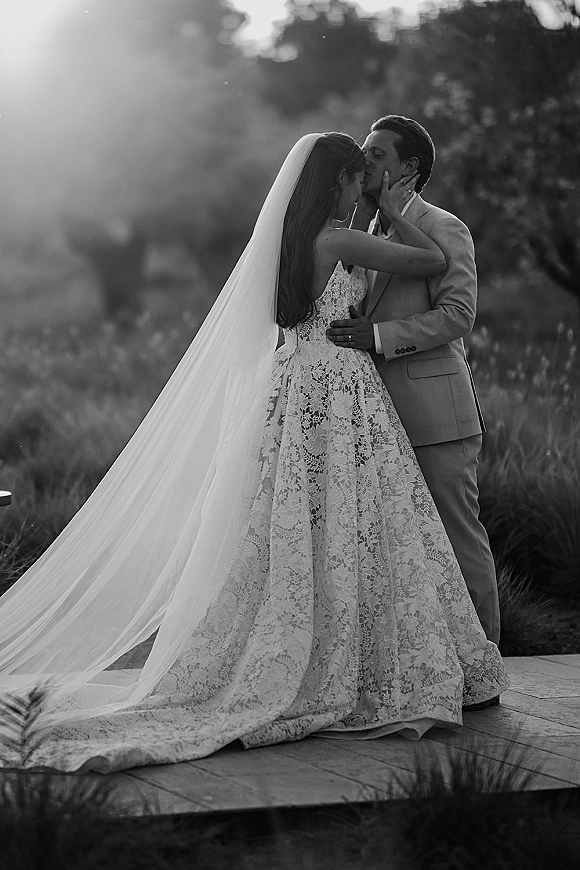 Wedding kiss portrait of bride and groom kissing, lace gown and long veil flowing, embracing on a stone patio with greenery behind