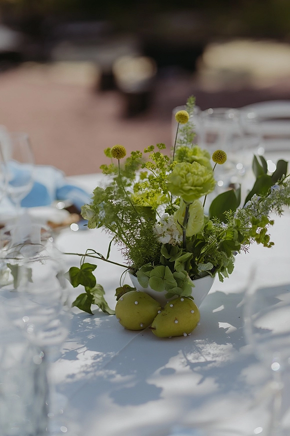 Wedding centerpiece with a green floral centerpiece in a white vase, lemons and pearl pins on a table set for an outdoor reception