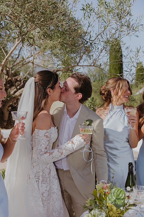 Wedding kiss as newlyweds hold champagne coupes, bride in lace dress and veil with groom in suit beneath olive branches and blue sky