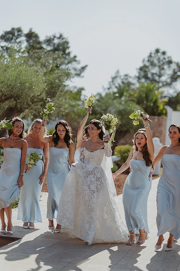 Bride with bridesmaids walking with bride, holding bouquets overhead, lace dress and long veil on a tree-lined outdoor walkway