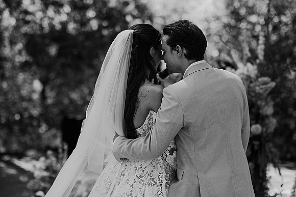 Wedding couple portrait in black and white, bride and groom embracing with foreheads touching, long veil and lace dress amid trees