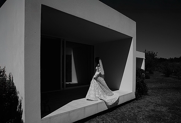 Bridal portrait of a bride in veil wearing a long sleeve lace gown with trailing train on a concrete terrace by modern windows