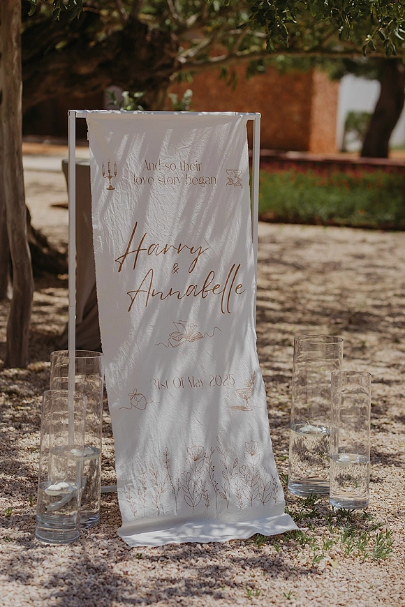 Wedding welcome sign with gold calligraphy on a fabric banner in a metal frame, styled with floating candle cylinders in a garden setting