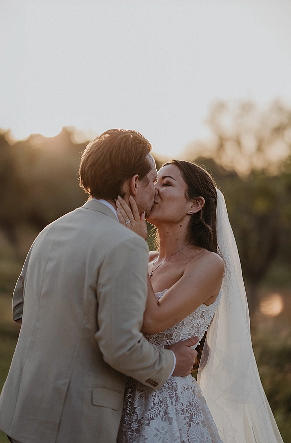 Wedding kiss portrait of bride and groom kissing as her veil blows, her hand on his face, backlit by sunset sky and trees