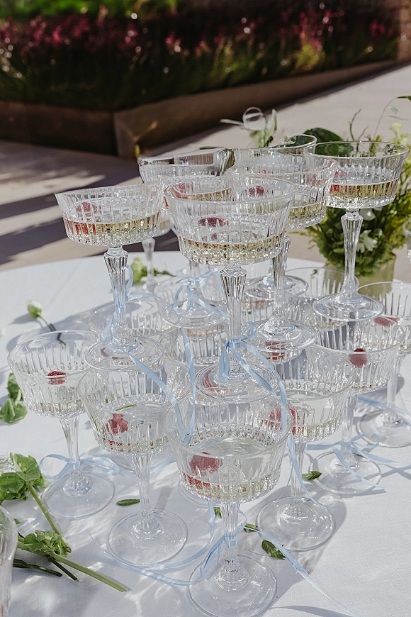 Champagne tower of coupe glasses filled with champagne and raspberries, tied with blue ribbon on a white tablecloth patio by a floral hedge