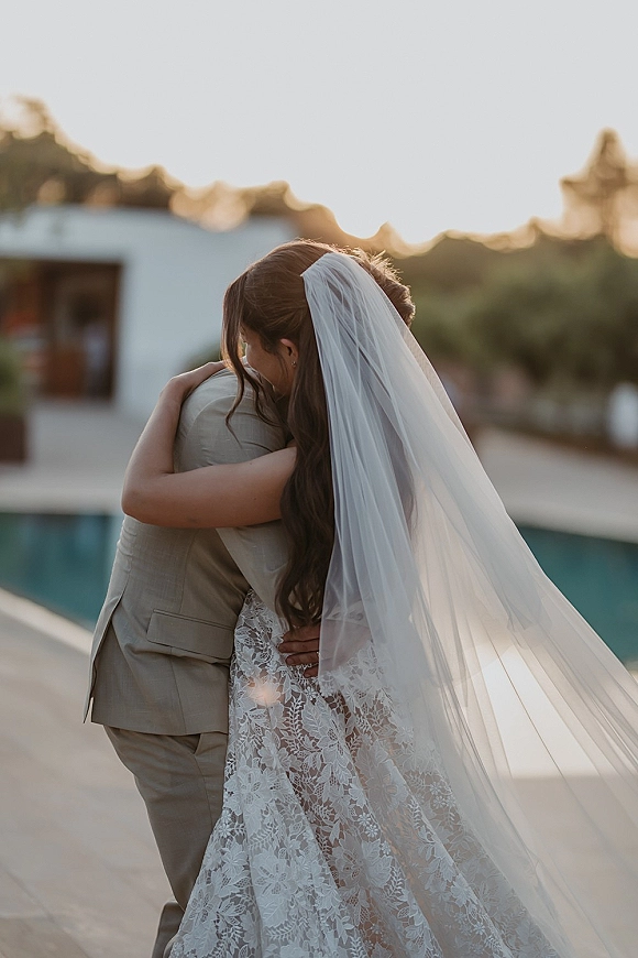 Couple portrait of bride and groom hug from behind, her long veil and lace dress in golden sunset by a poolside patio and modern villa