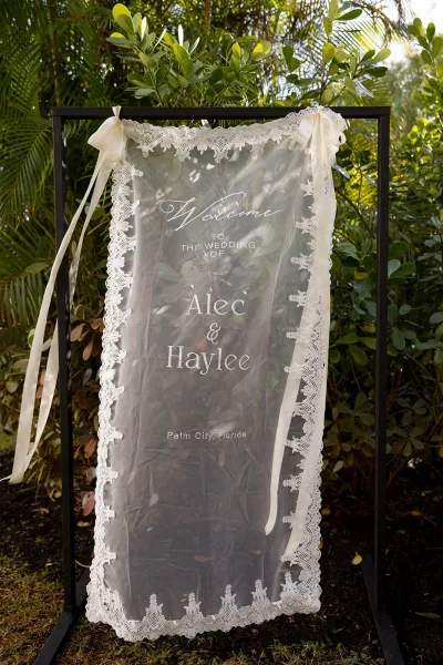 Wedding welcome sign on clear acrylic with calligraphy lettering, ivory ribbons, and lace trim in a black frame amid tropical greenery