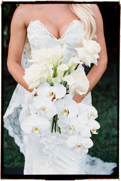 Bridal bouquet with a white orchid bouquet cascading with calla lilies and roses, held by a bride in a lace dress amid garden greenery
