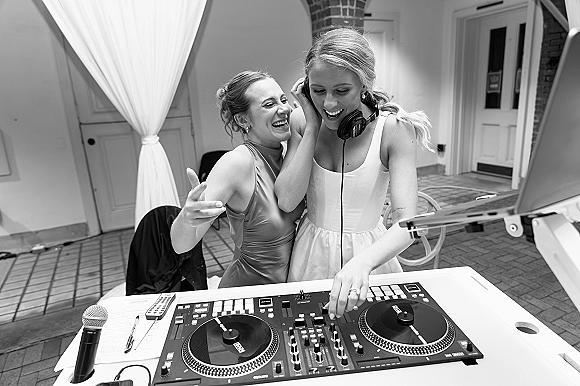 Wedding DJ moment as bride wearing headphones laughs with a bridesmaid at a DJ table with controller, mic and laptop on patio pavers