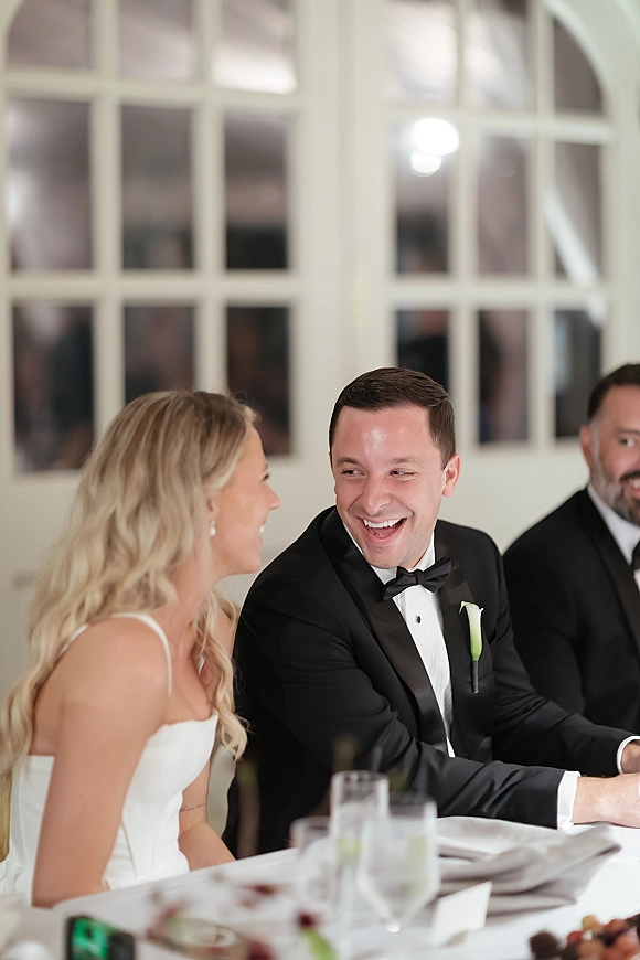 Wedding reception moment as bride and groom laughing at their head table, toasting with a champagne flute in an indoor room with white paned windows