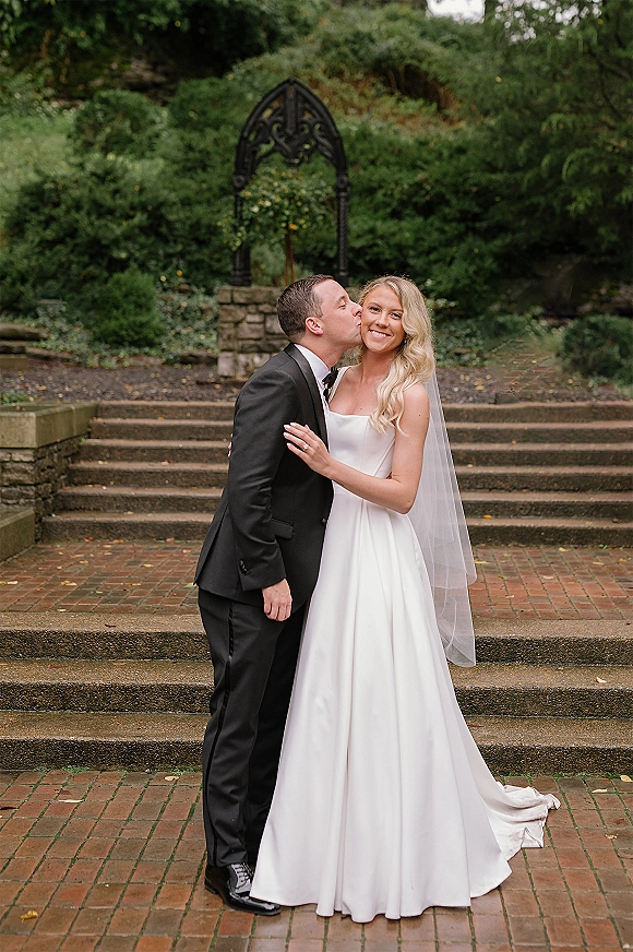 Wedding couple portrait of bride and groom kiss on stone steps, her long veil and satin dress flowing beside his black bow tie tuxedo under a wrought iron arch