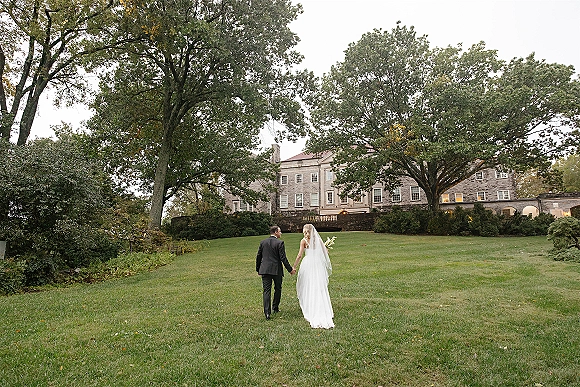 Couple portrait of bride and groom walking away hand in hand, her cathedral veil and calla lily bouquet on manor lawn under overcast sky