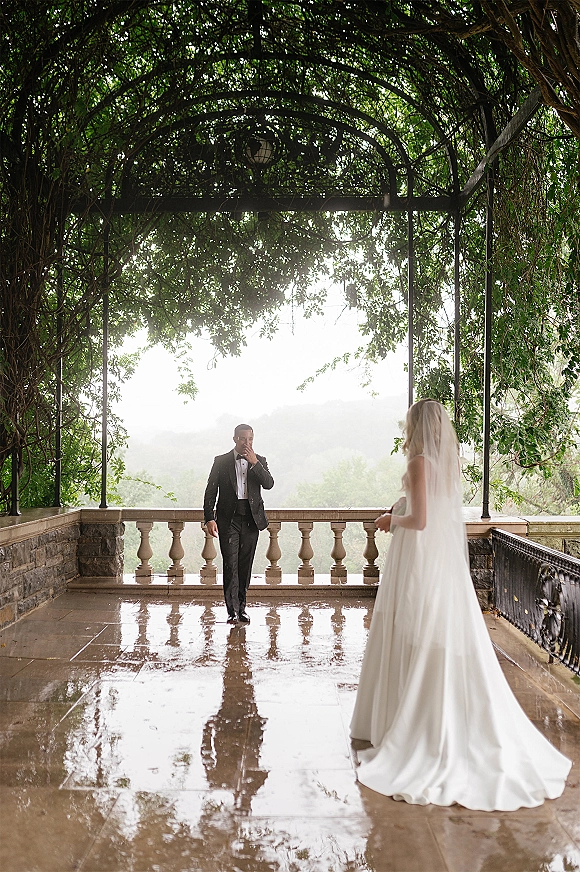 First look moment as bride in strapless gown and long veil approaches groom in tuxedo under vine-covered pergola on wet terrace