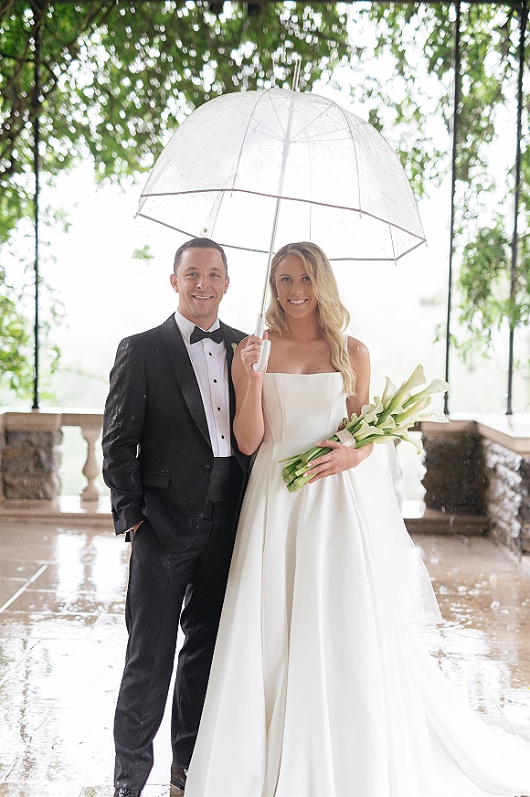 Wedding couple portrait of bride and groom under a clear umbrella on a rainy terrace, she holds calla lilies by a stone balustrade lake view