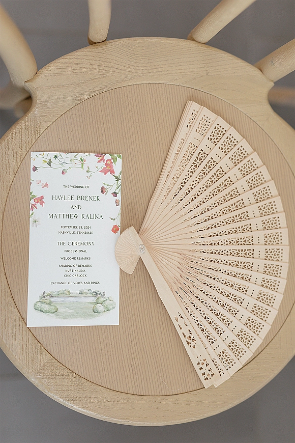 Wedding program and ceremony program template card with floral border beside a laser-cut wooden hand fan on a chair seat