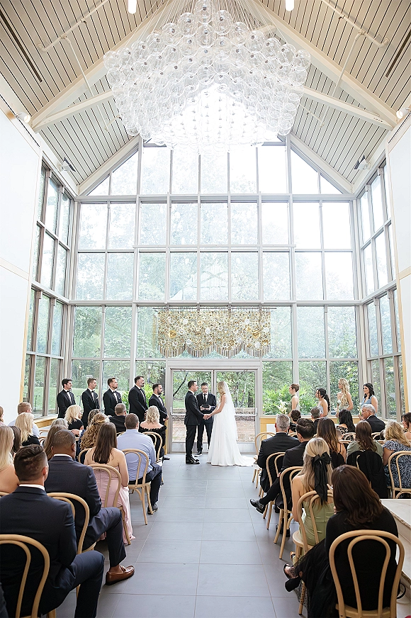 Wedding ceremony wide shot with bride and groom at altar, glass chandelier overhead, guests seated in rows by floor-to-ceiling windows